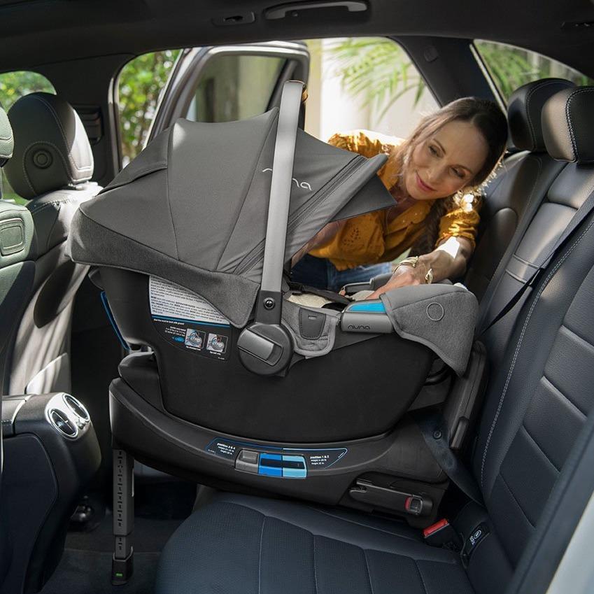 Rear-facing infant car seat with its canopy up, installed into back seat of car, mom is leaning into the car to check that baby's harness is secure.